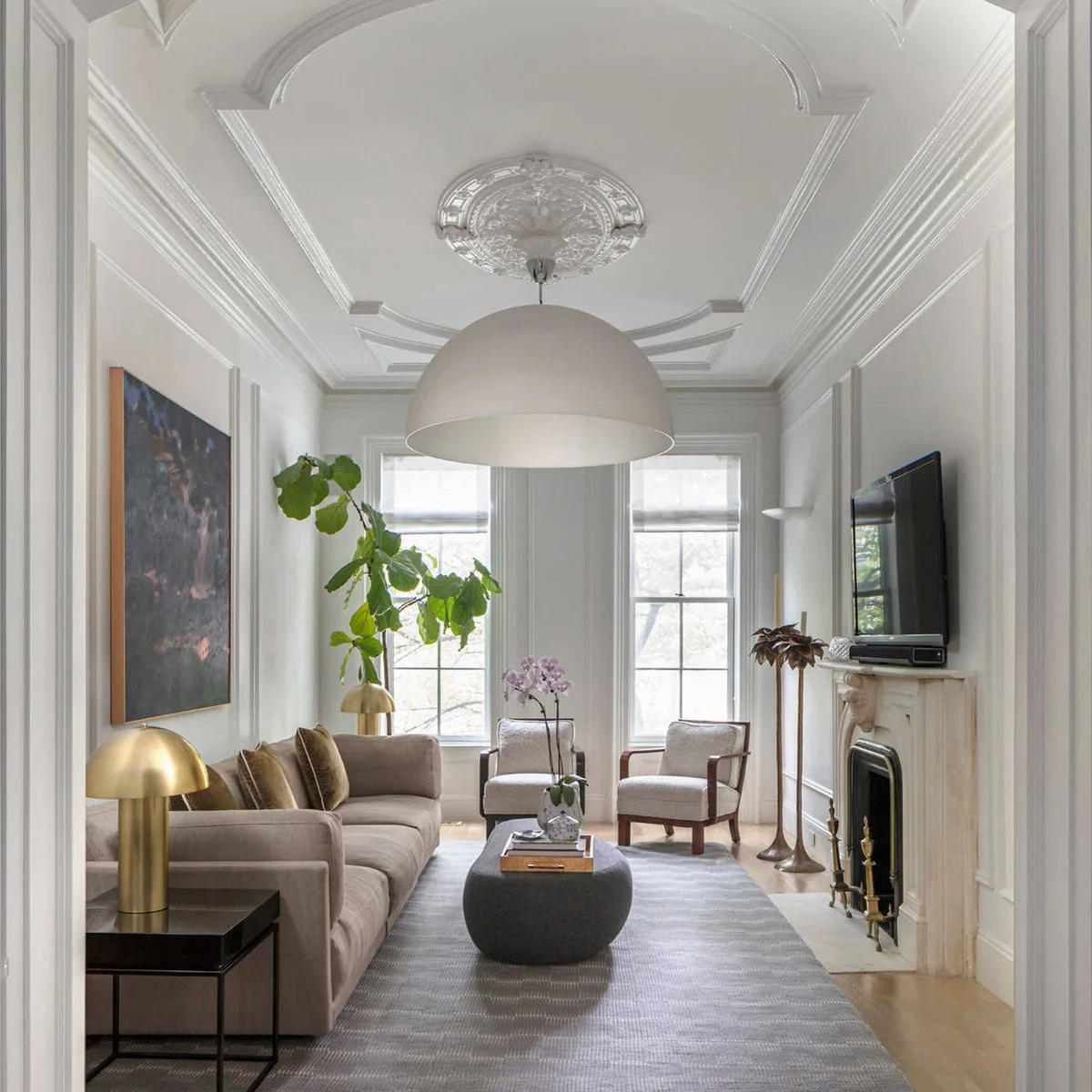 Narrow white living room with a taupe sofa, two wood-framed chairs, oval ottoman and fireplace opposite