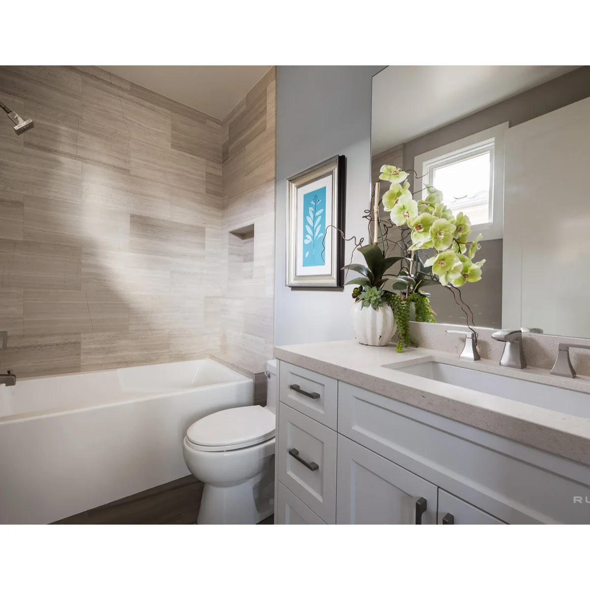 Small bathroom with a grey vanity, broad mirror and a white tub below pale horizontal wall tiles