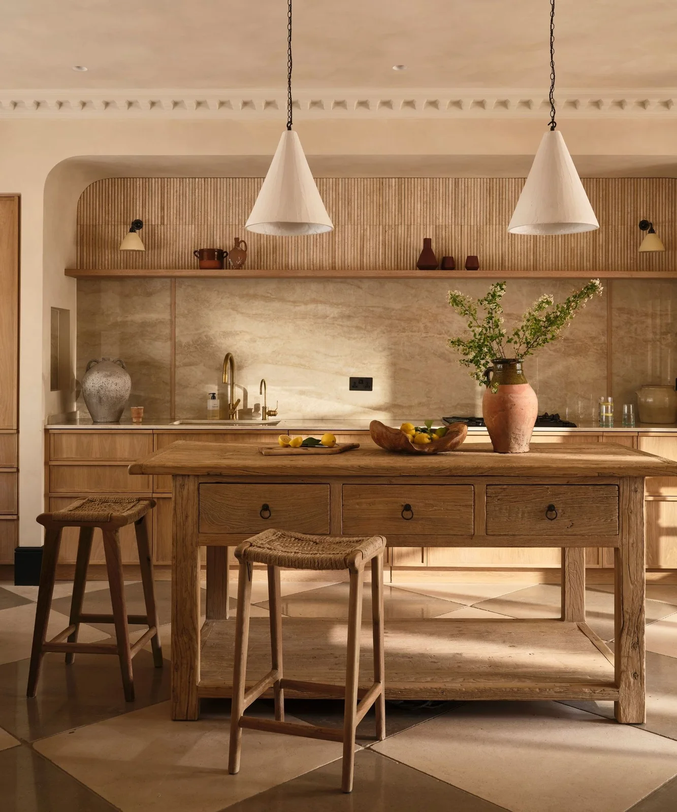 A wooden kitchen island and woven stools beneath pale cone pendants in a warm neutral room