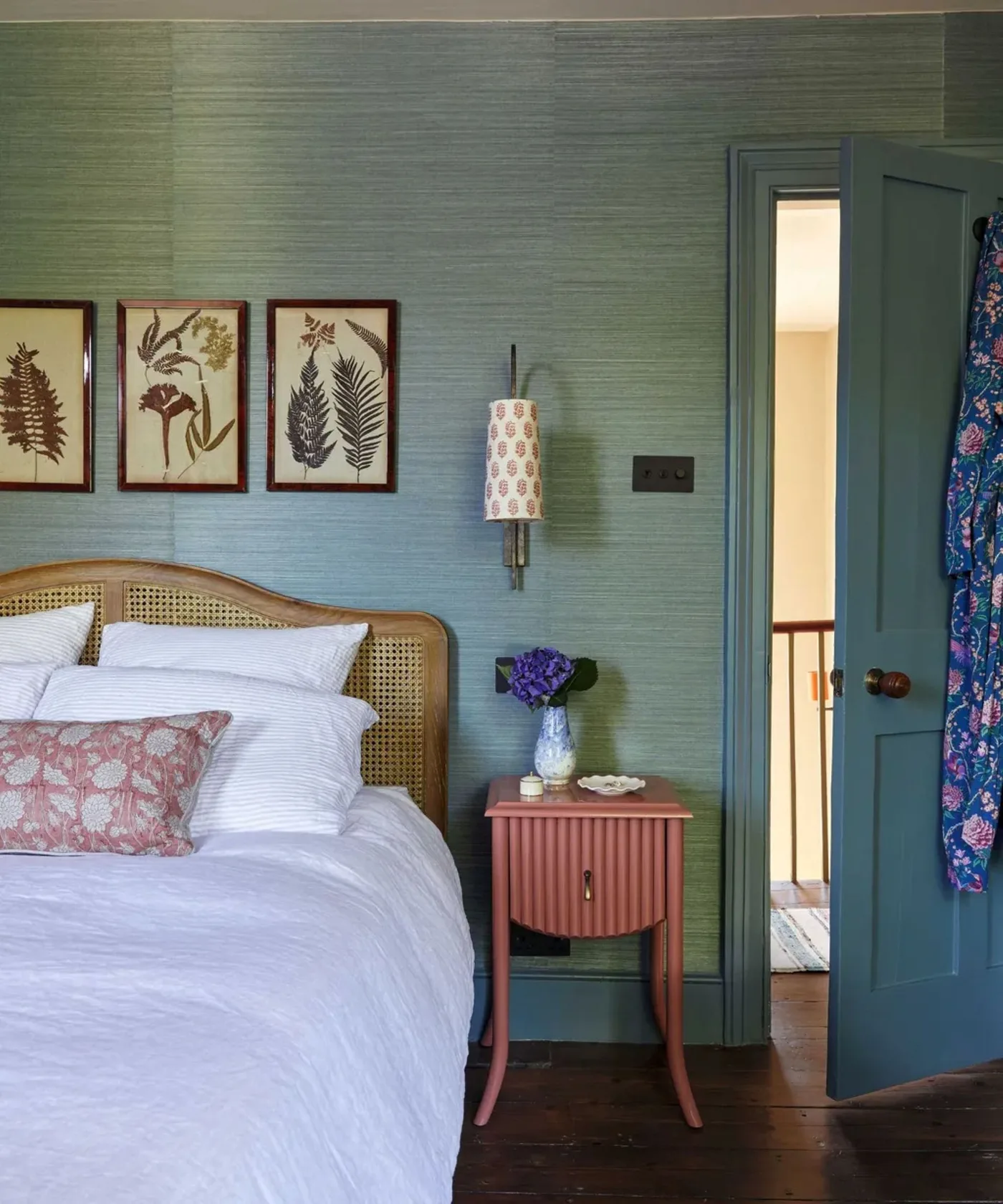 Blue-green textured wallpaper behind a woven headboard, botanical prints, a pink bedside table and a teal door.
