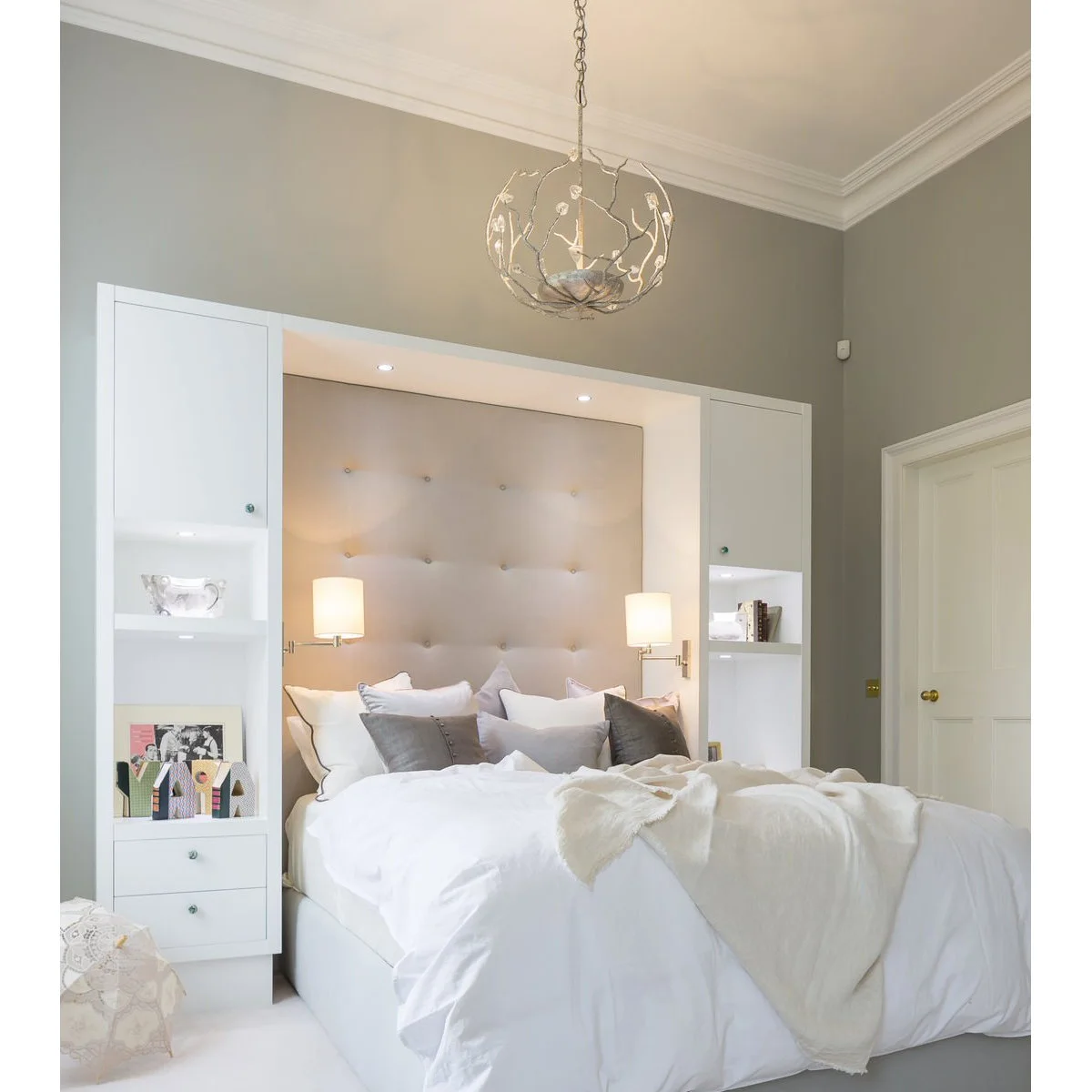 Bed with pale padded headboard framed by white cupboards, shelves and two wall lights