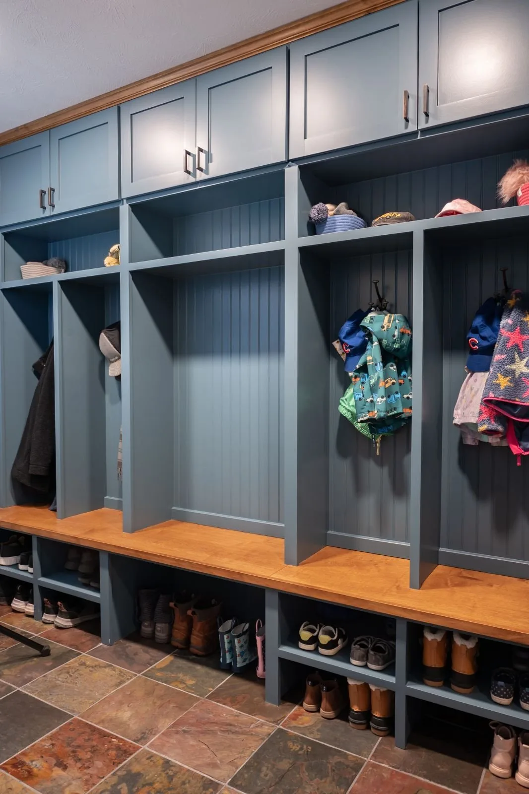 Blue built-in mudroom bays with a long wooden bench, upper cabinets, hanging coats and shoe shelves over multicolored tile.