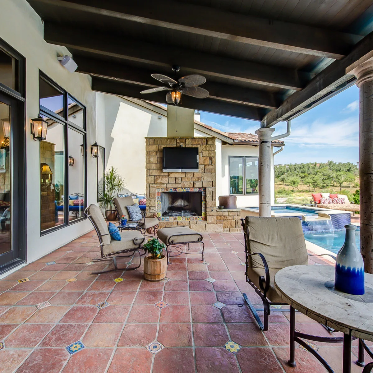 Covered patio with warm floor tiles and small patterned inserts beside a stone fireplace, pale chairs and blue cushions.