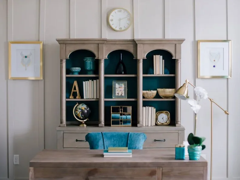 Gray office cabinet with arched shelves and a turquoise chair behind a wooden desk with warm metal lamps.