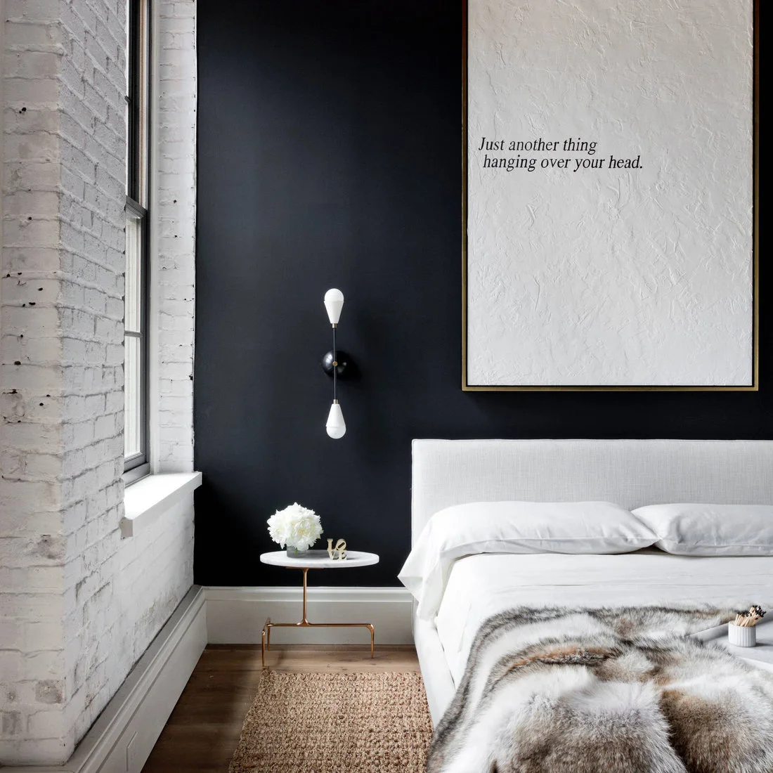 Large pale typographic artwork above a white bed against a black wall beside painted brickwork.