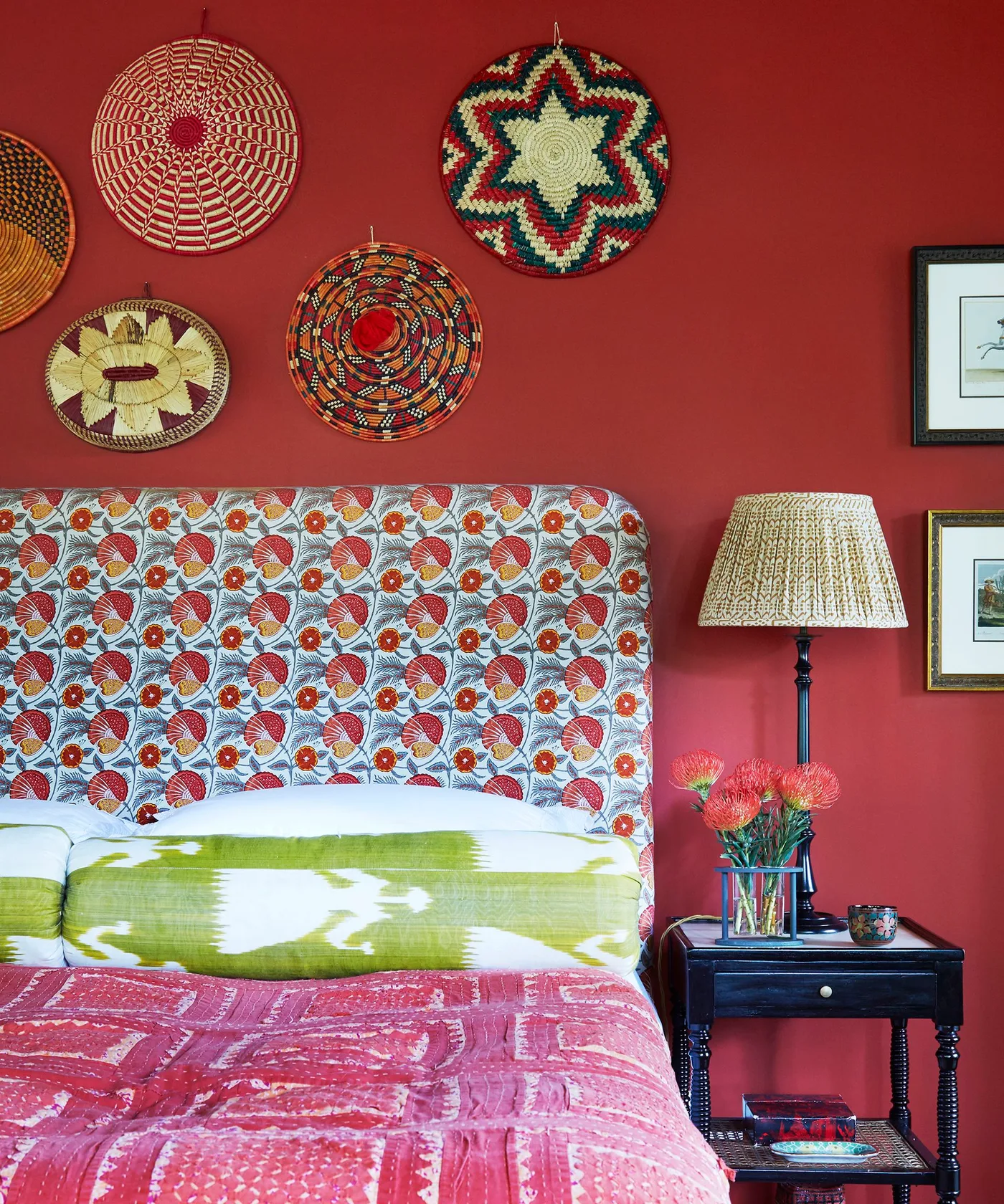 Rich red bedroom wall with decorative woven plates, floral upholstered headboard, patterned bedding, and a woven lampshade.