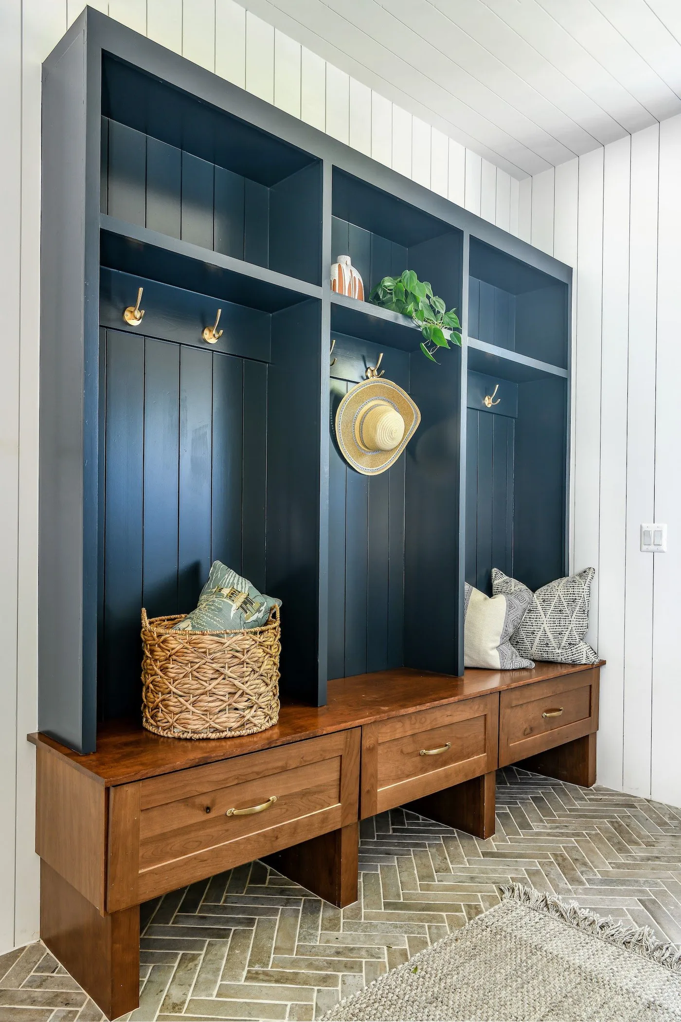 Navy mudroom hanging bays with upper shelves, gold-tone hooks, wood bench and drawers, woven basket, white paneled walls, and herringbone-patterned floor.
