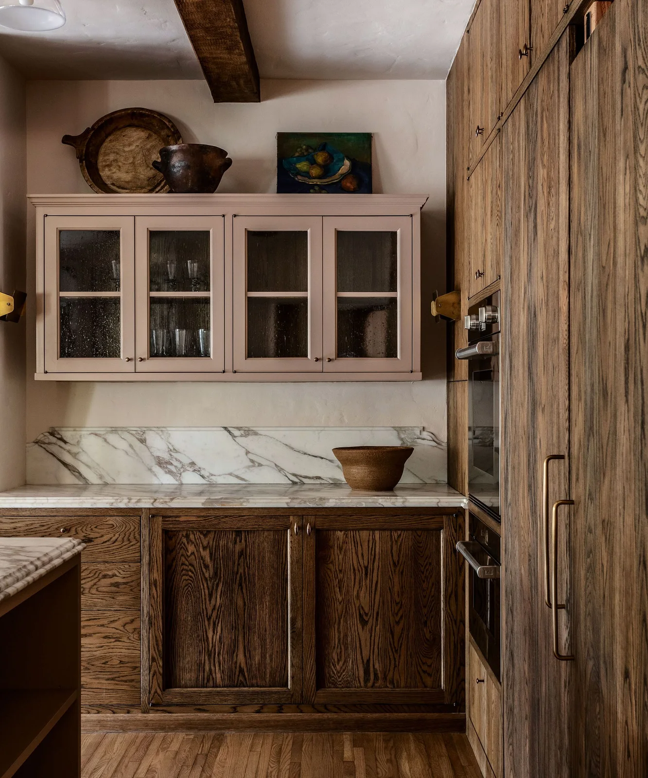 A still-life picture, broad handled tray and dark rounded pot above a pale glass-front kitchen cabinet beside wood-grain cabinetry.