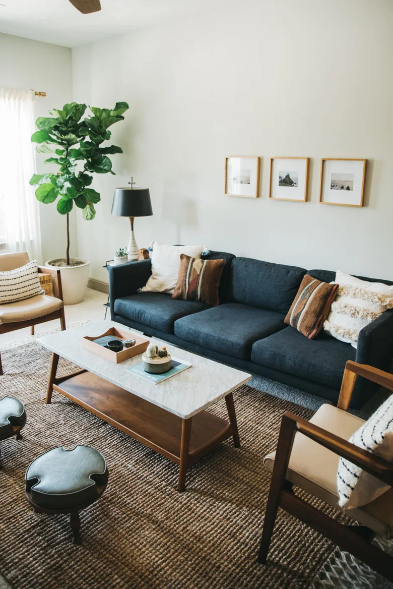 A dark blue sofa stands against a pale wall with three small framed pictures, striped cushions, wood armchairs and a pale-topped wood coffee table.
