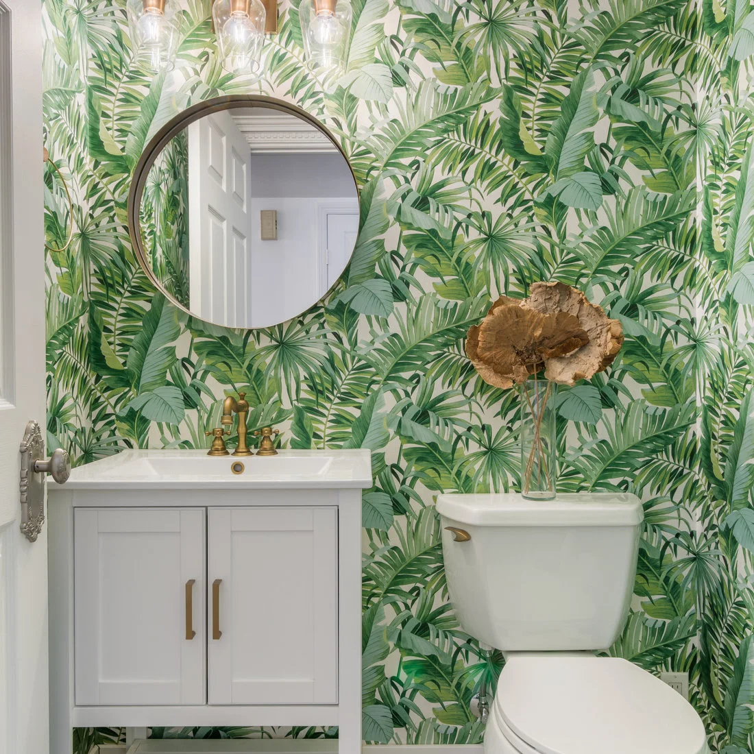 Dense tropical leaf wallpaper surrounds a round powder-room mirror above a pale gray vanity beside gold-colored fittings and a white toilet