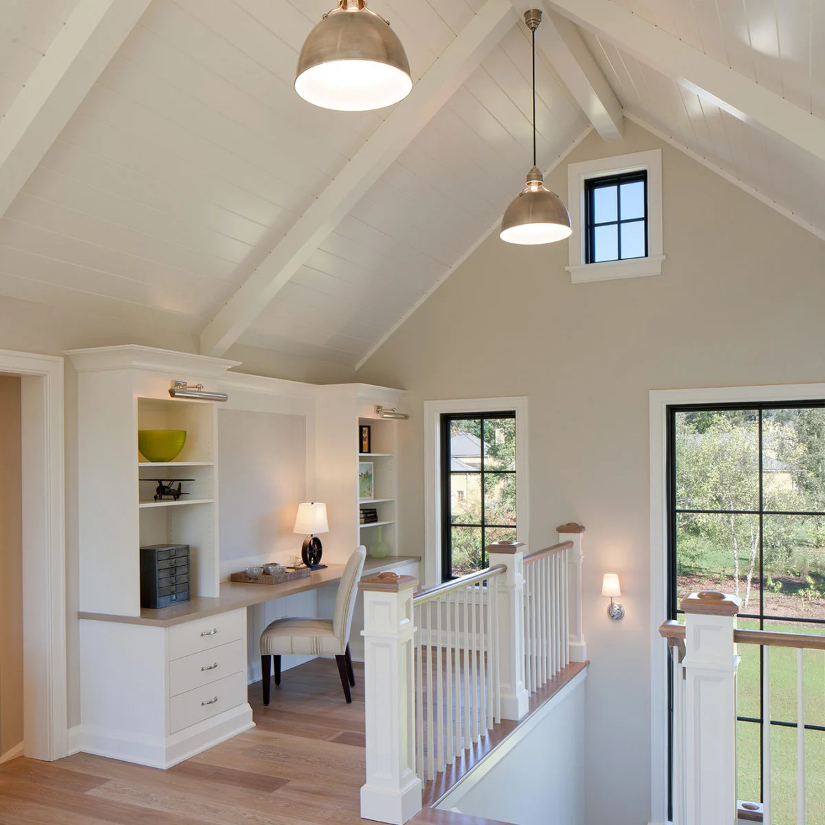Landing home office with pale built-in desk, shelves, upholstered chair and white railing beneath a vaulted ceiling
