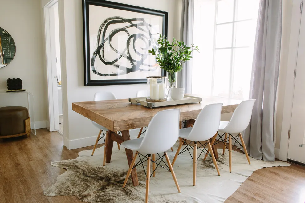 A timber table with white chairs sits beneath artwork beside a broad window