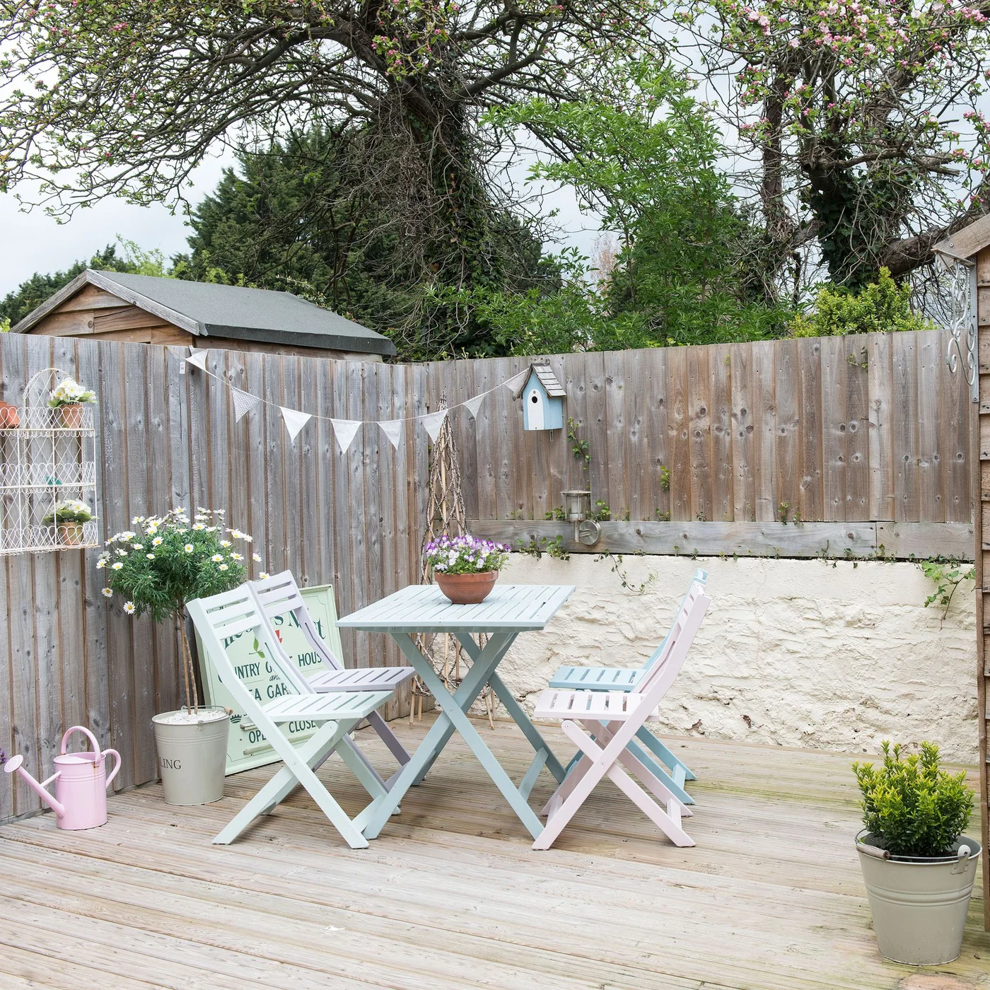 Pastel folding chairs and a small table sit on timber decking beside a fenced garden corner
