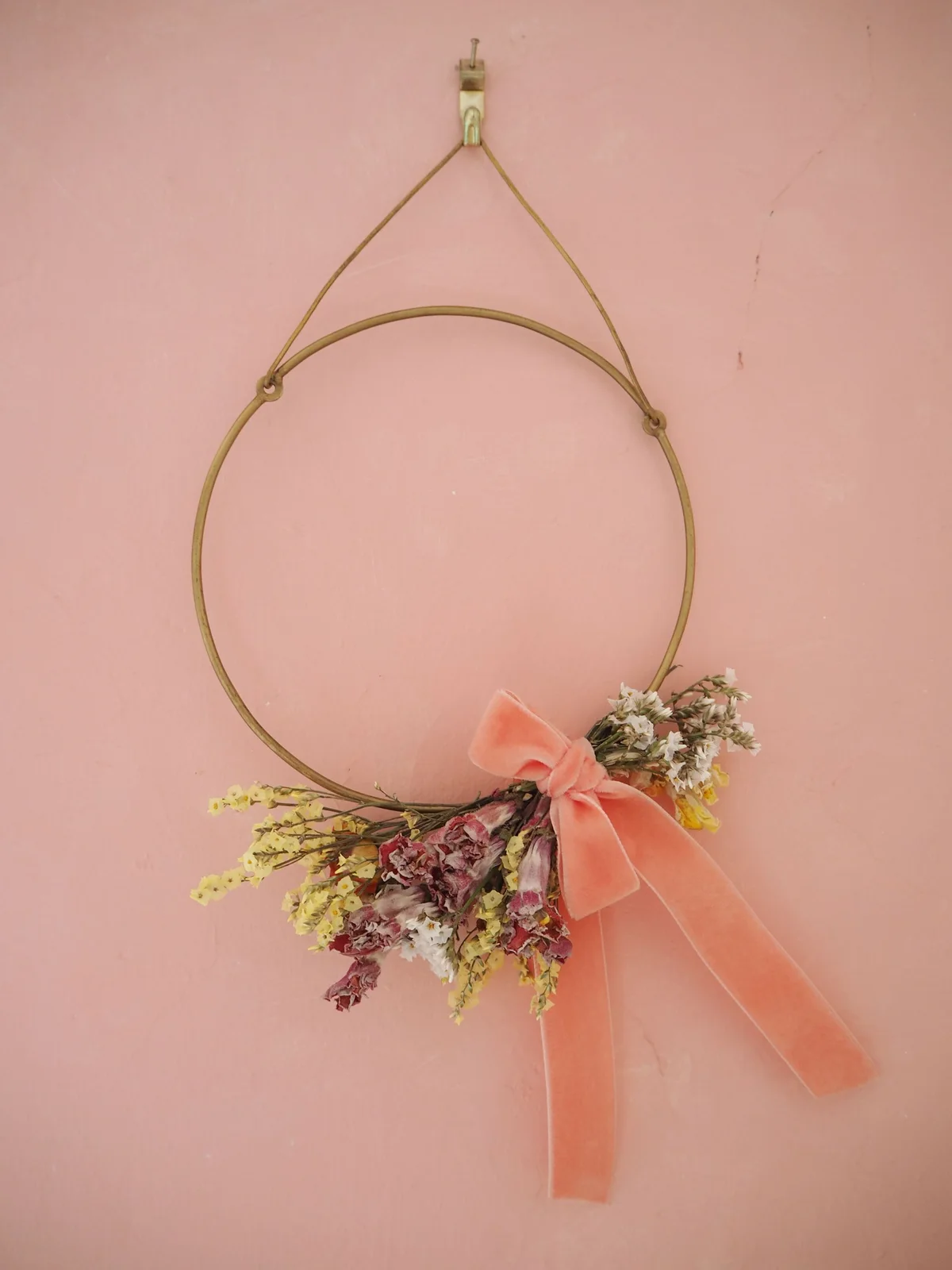 A thin circular hoop carries a small dried flower cluster and pink ribbon at its lower edge against a pink wall.