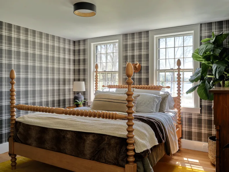 A turned timber four-poster bed stands against checked walls beside large windows and neutral bedding.