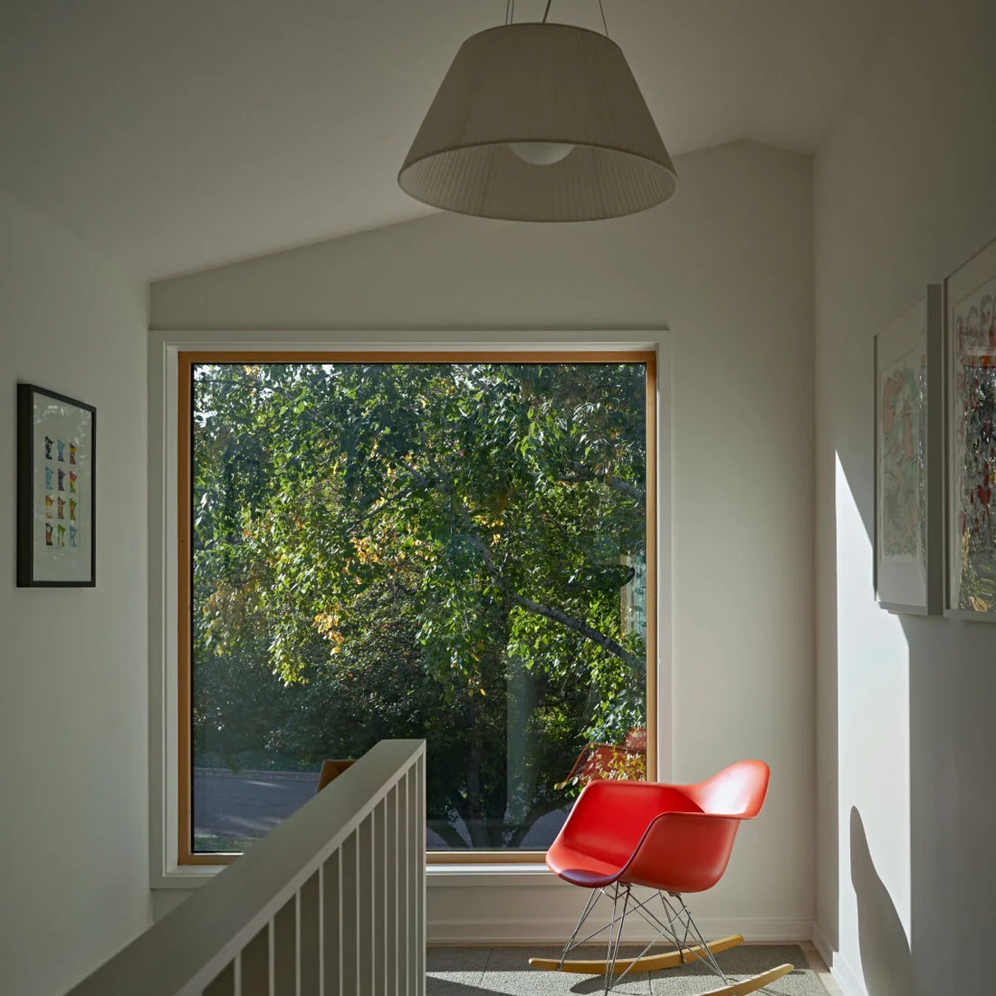 A red rocking chair stands beside a broad picture window on a white stair landing with framed art and a pale rug.