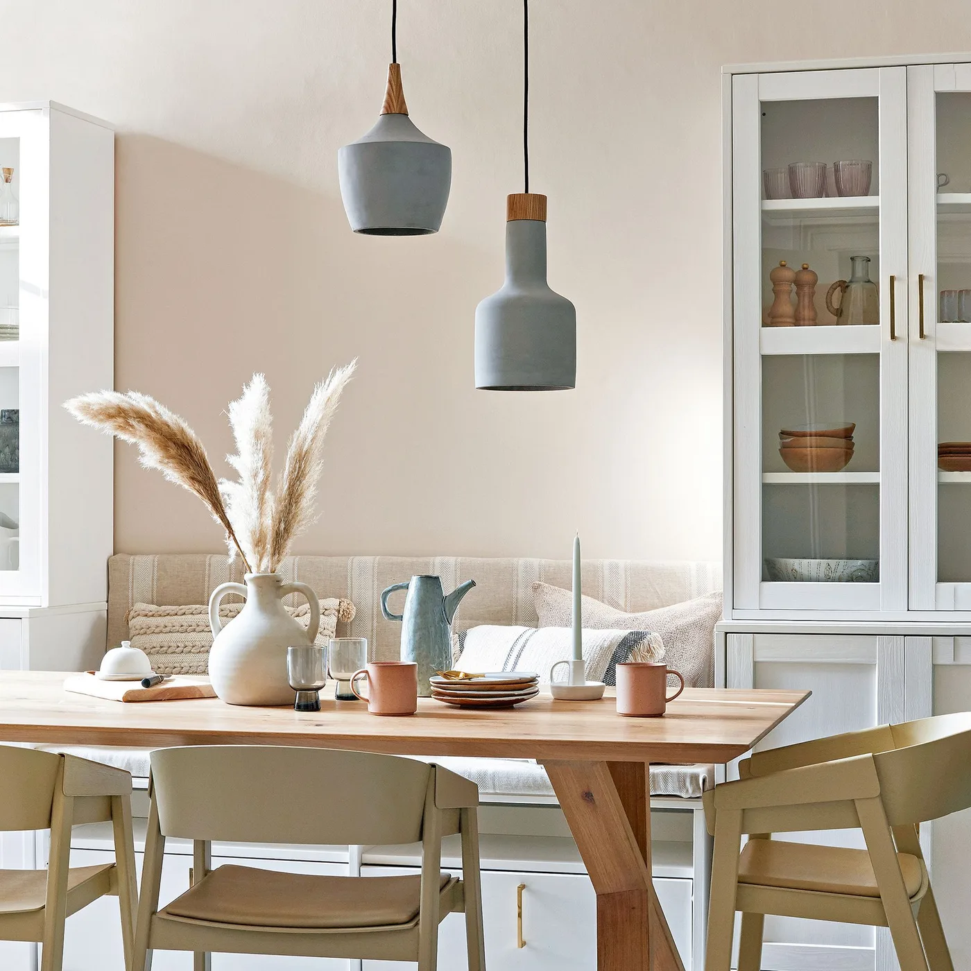 Country dining room with pale cupboards around a cushioned bench, wooden table, painted chairs and two blue-gray hanging lights.