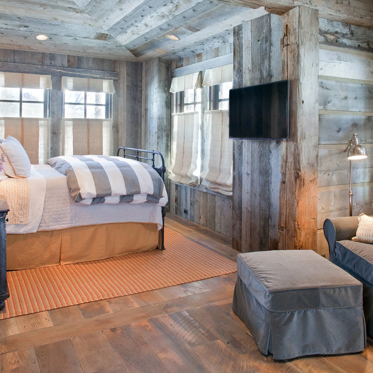 Wood-lined bedroom with striped bedding, pale window shades and blue chair with ottoman