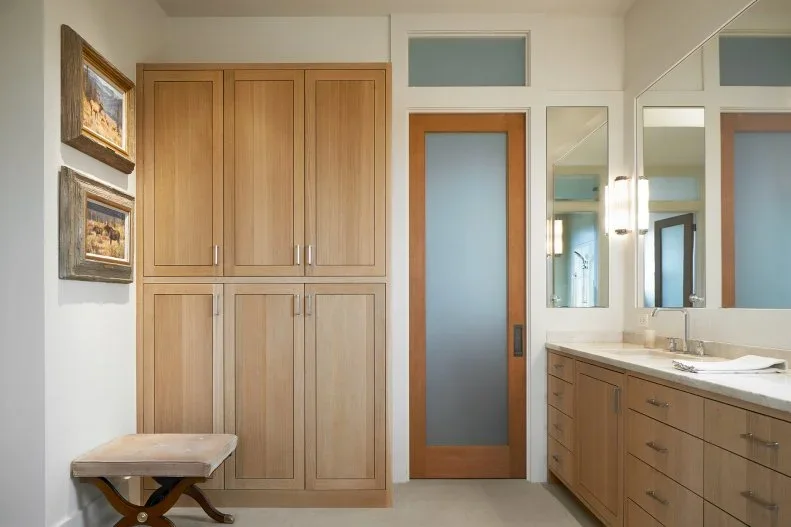 Tall wood cupboards and lower drawers stand beside a long pale bathroom counter, wall mirrors and a wood-framed frosted door