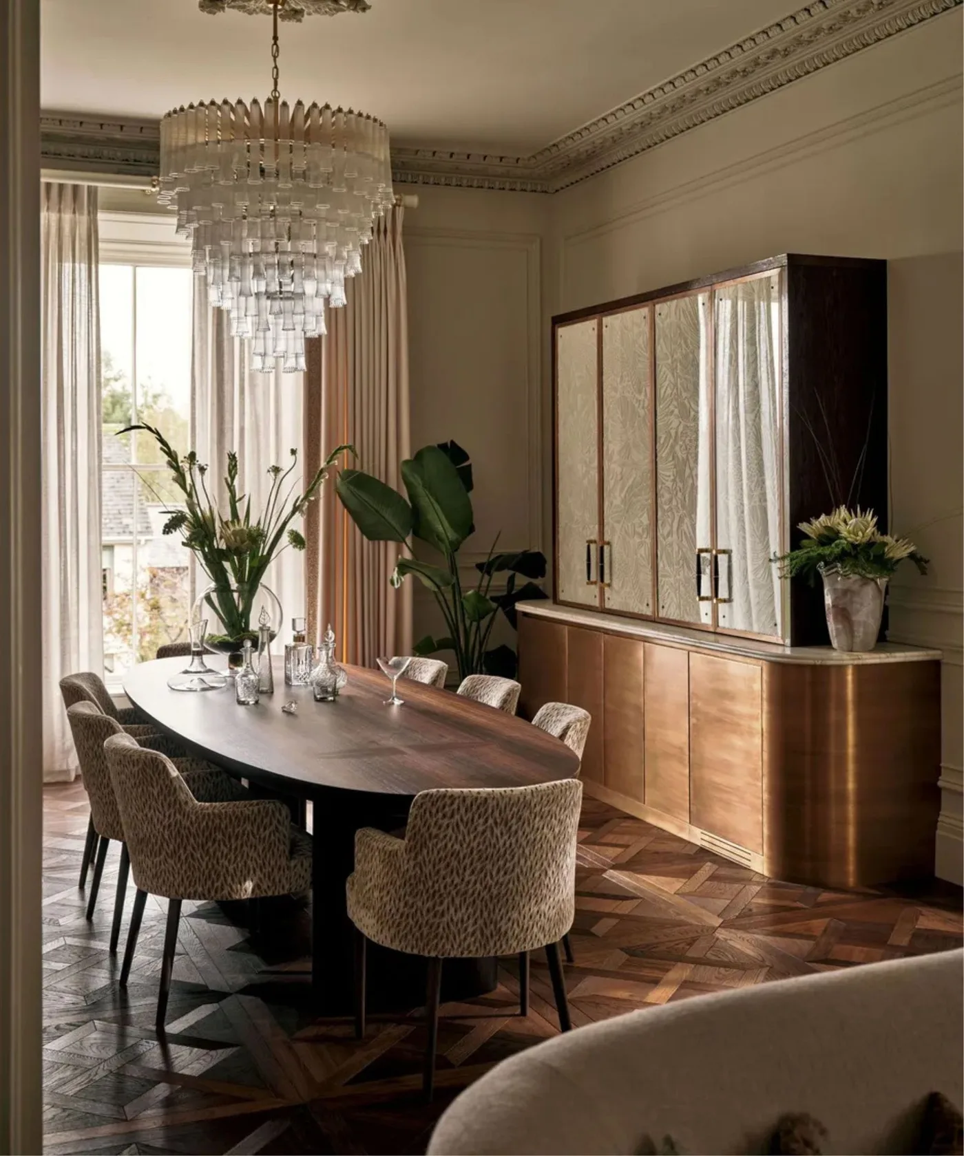 A tiered hanging light sits above an oval dining table and upholstered chairs beside a pale cabinet and tall curtains