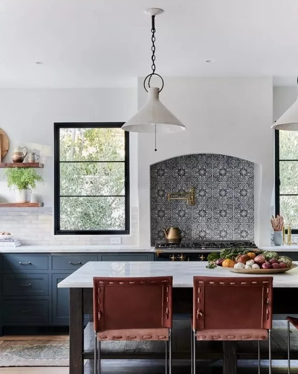 A blue kitchen has patterned tiles within a curved cooking recess between dark-framed windows beside a pale island, warm brown stools and white pendants