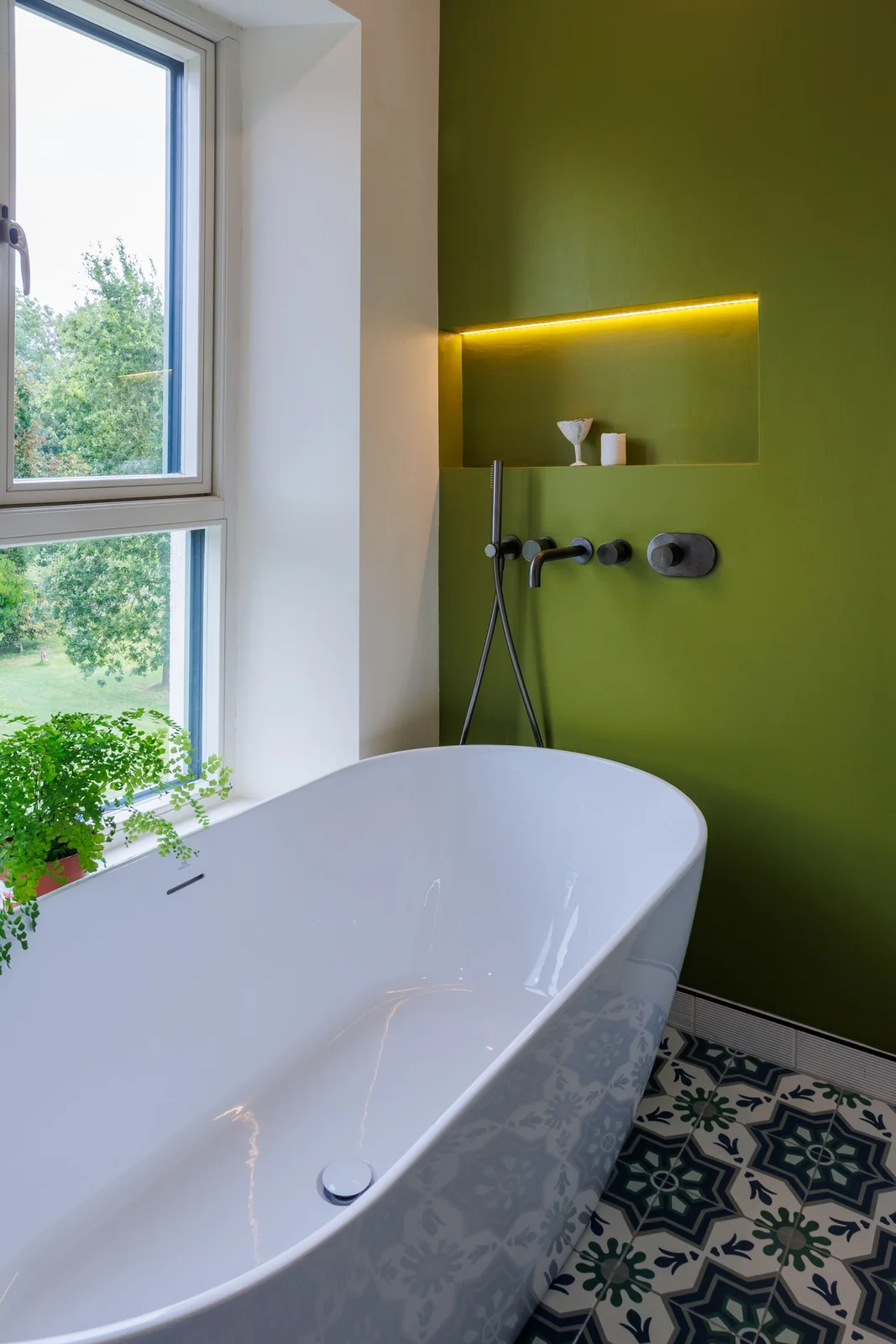 A white freestanding bath sits against an olive wall with a lit recessed niche, black wall-mounted fittings and a patterned floor.