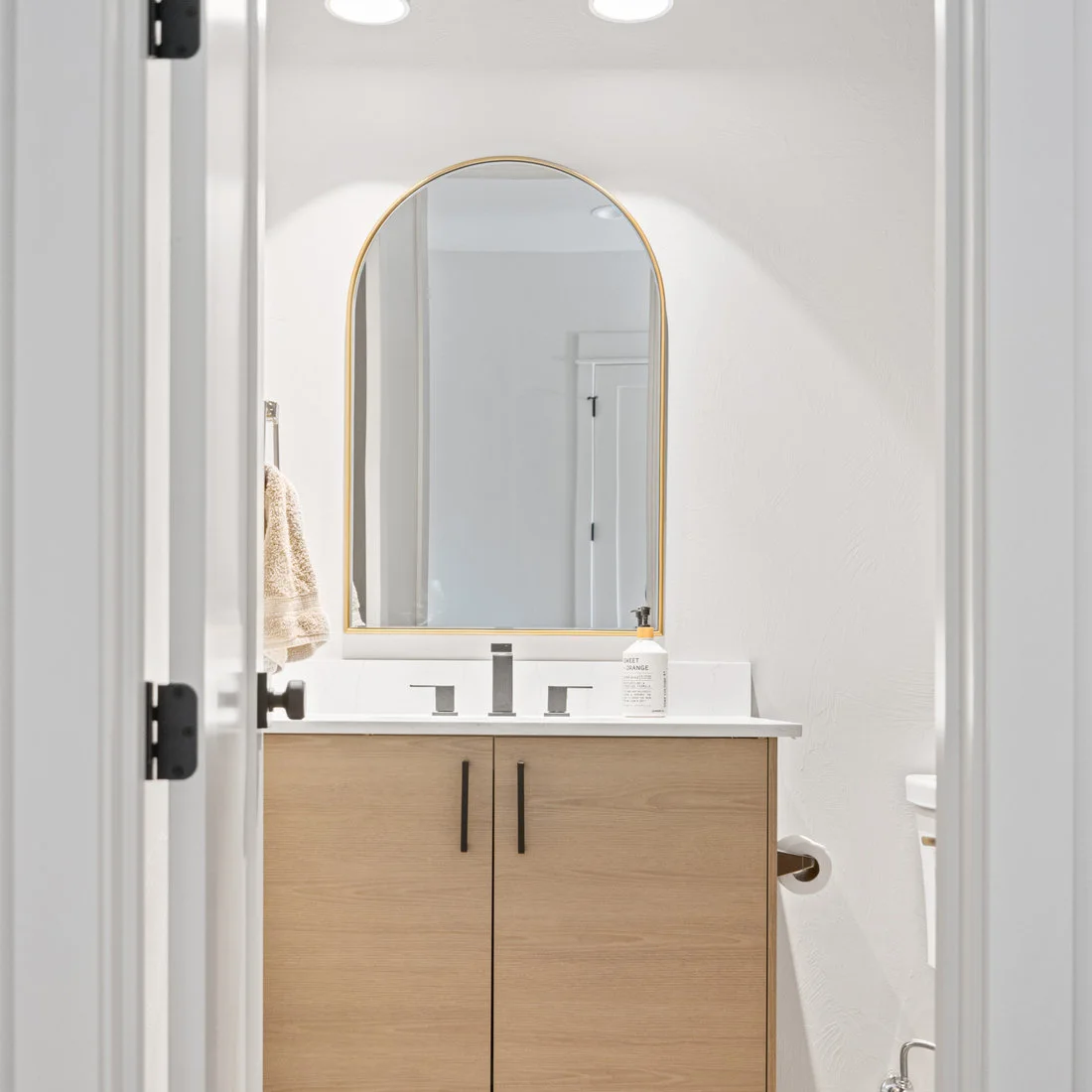 A pale wood two-door vanity with black handles and white counter sits beneath an arched warm-metal mirror against plain white walls.