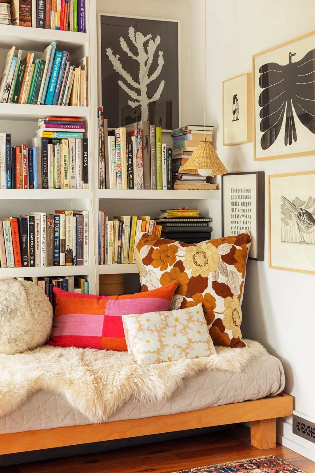 A cushioned wooden bench with colorful pillows sits beside white bookshelves and botanical artwork beneath a small woven-shade lamp