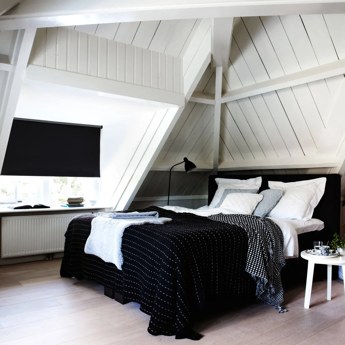 Attic bedroom with white painted roof boards, a black window blind and dark bedding on a low bed.