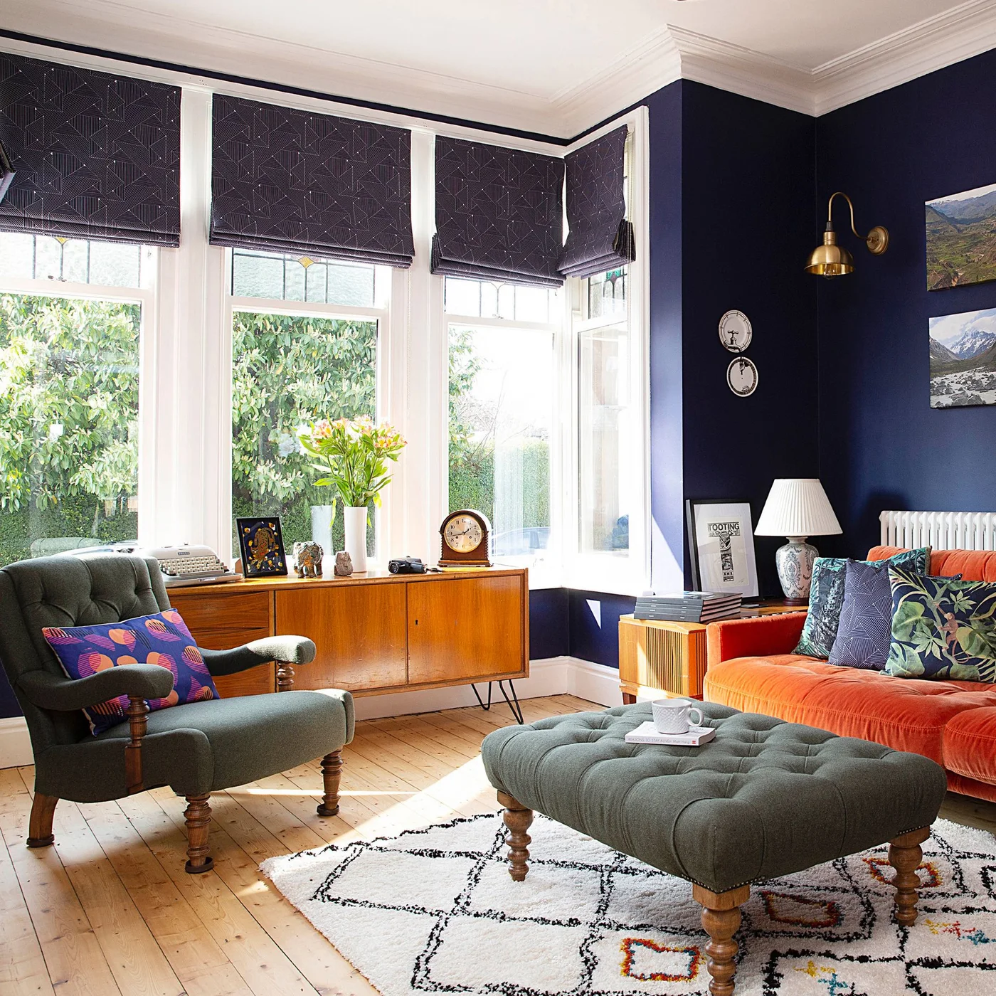 An orange sofa and green armchair sit beside a wooden sideboard beneath dark blue walls and patterned blinds