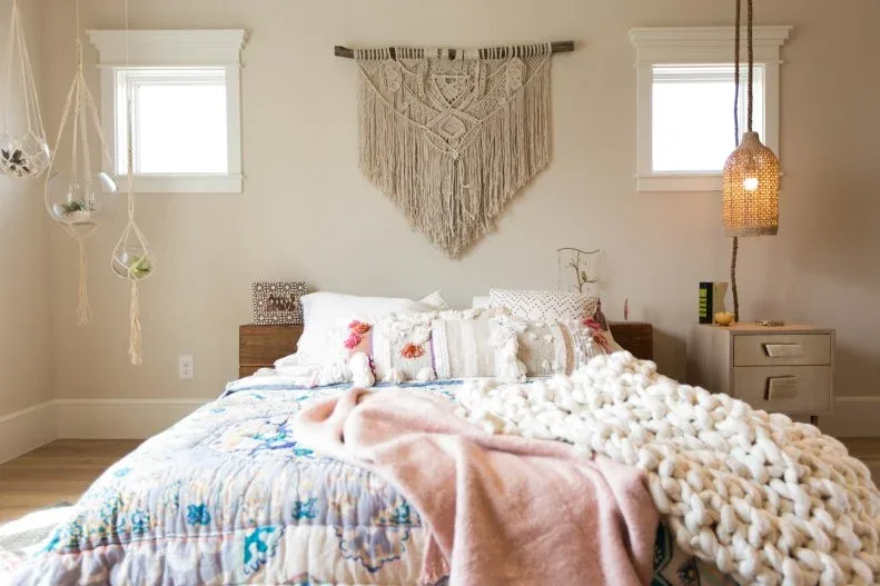 A fringed woven hanging sits between small upper windows above layered pale bedding with blue and pink details beside a small chest and woven pendant