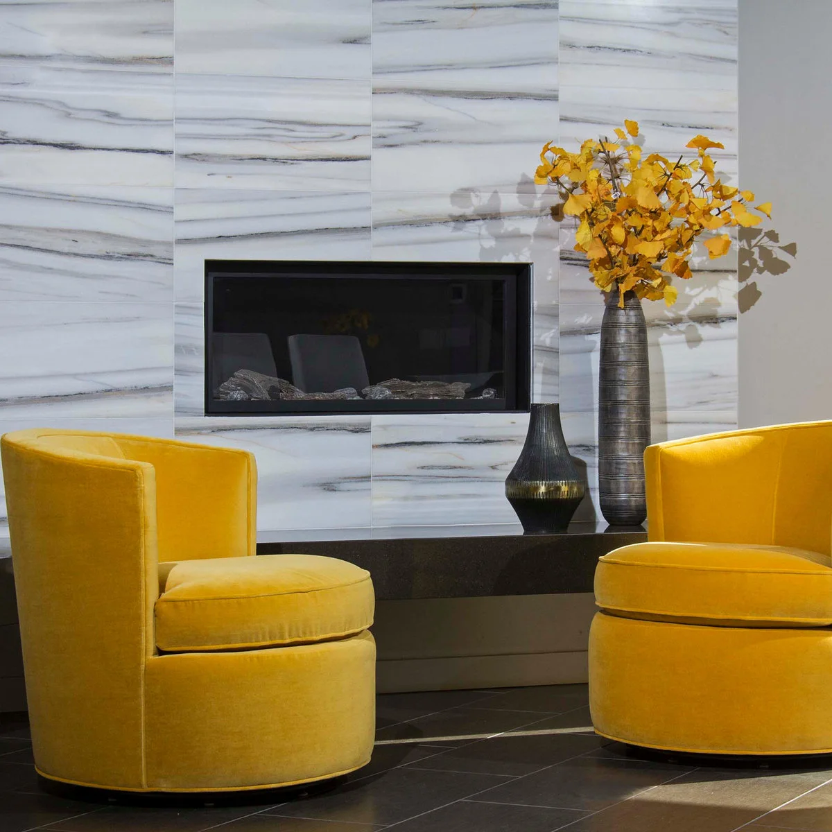 Two mustard yellow chairs face a pale gray veined fireplace wall above dark gray floor tiles.
