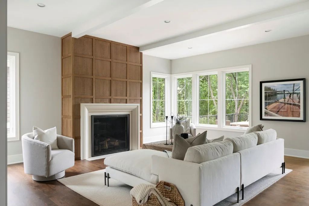 Living room with a wood-paneled fireplace wall, pale sectional sofa, rounded armchair and a large garden-facing window.