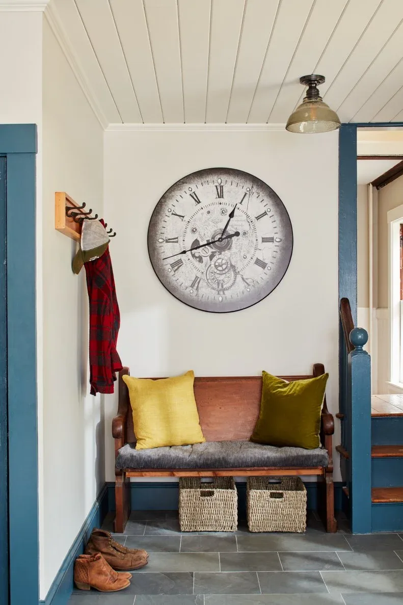 Small entryway with wooden bench, woven baskets below, blue door trim, pale boarded ceiling and large round wall clock.
