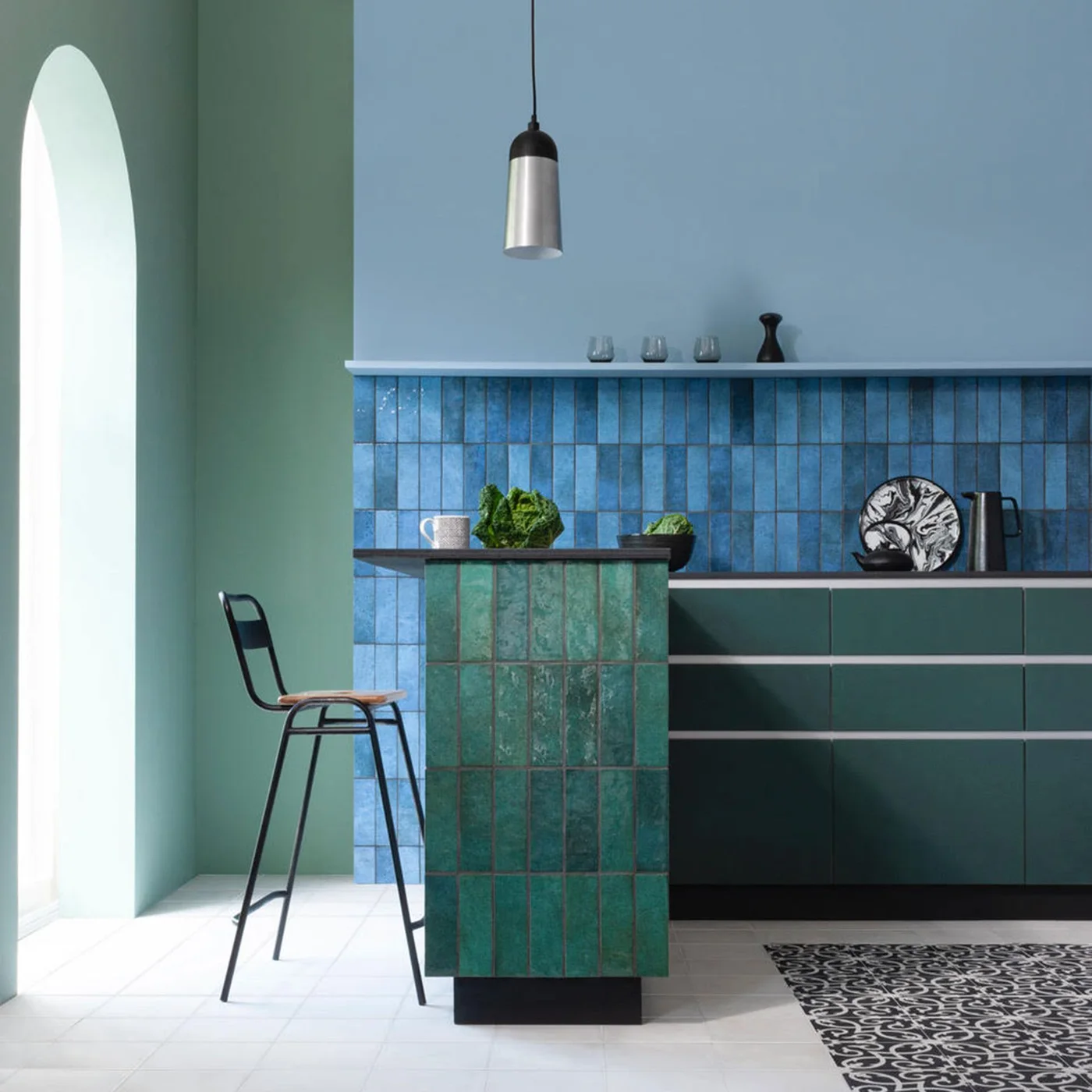 Blue backsplash tiles and green tiles on a narrow kitchen peninsula sit beside dark green cabinets and pale flooring