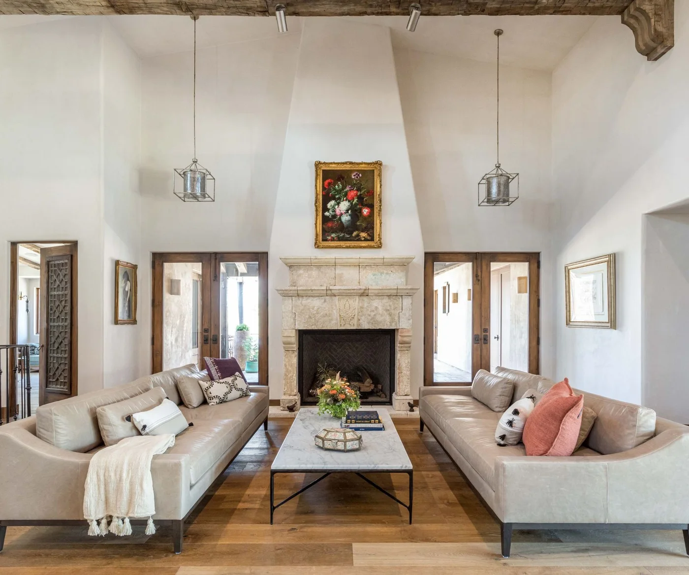 Two pale living-room sofas face each other beside a tall fireplace wall, wooden glazed doors, hanging lantern-like lights and a slim coffee table