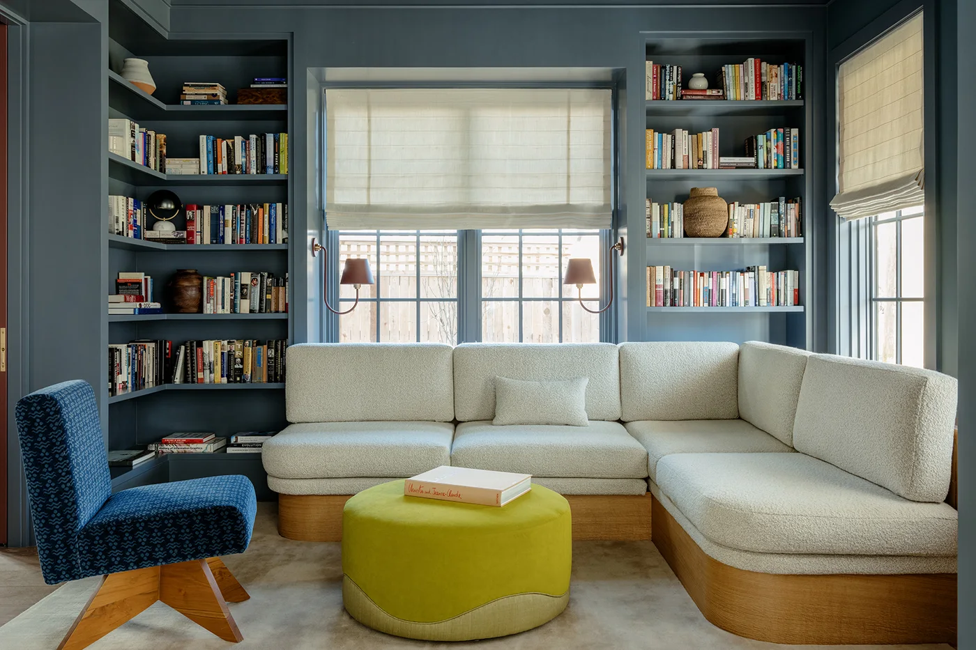 A pale wraparound sofa sits between blue bookcases and windows beside a blue chair and bright yellow-green ottoman