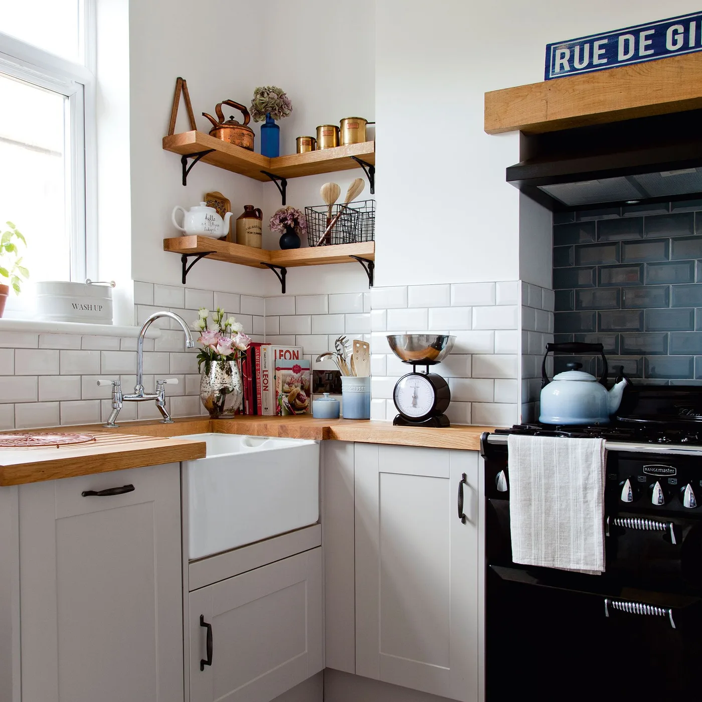 Small white framed kitchen with an apron-front sink, wooden worktops, open corner shelves and a dark tiled cooker alcove.