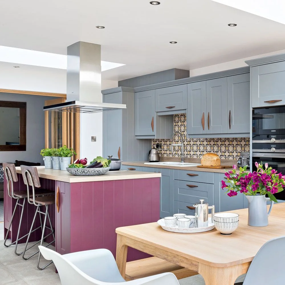 A plum-colored island stands between gray kitchen cabinets and a wooden dining table beneath a metal extractor