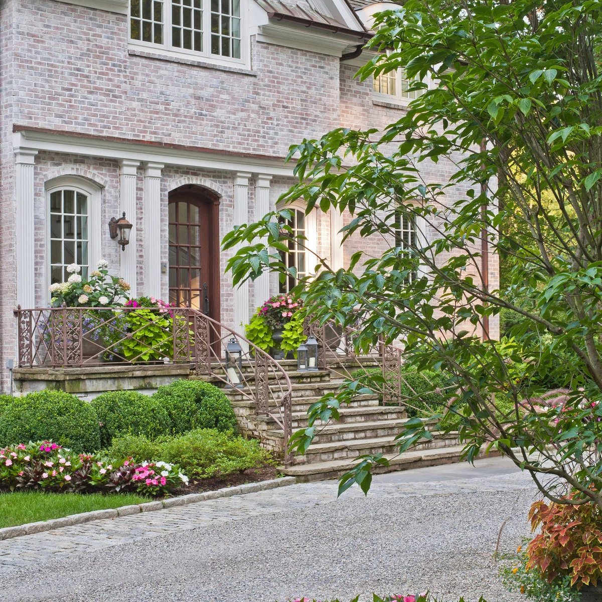Gravel driveway beside broad front steps, stone edging, clipped shrubs and large flower-filled porch containers.