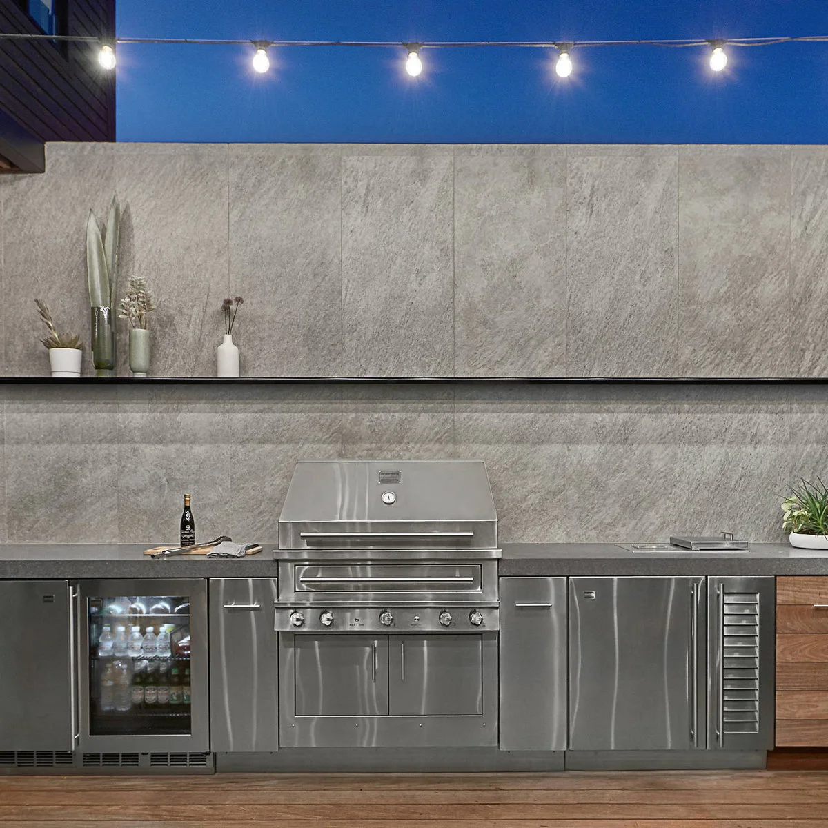 Outdoor kitchen with a central grill, gray backsplash, metal cabinets, beverage refrigerator and string lights