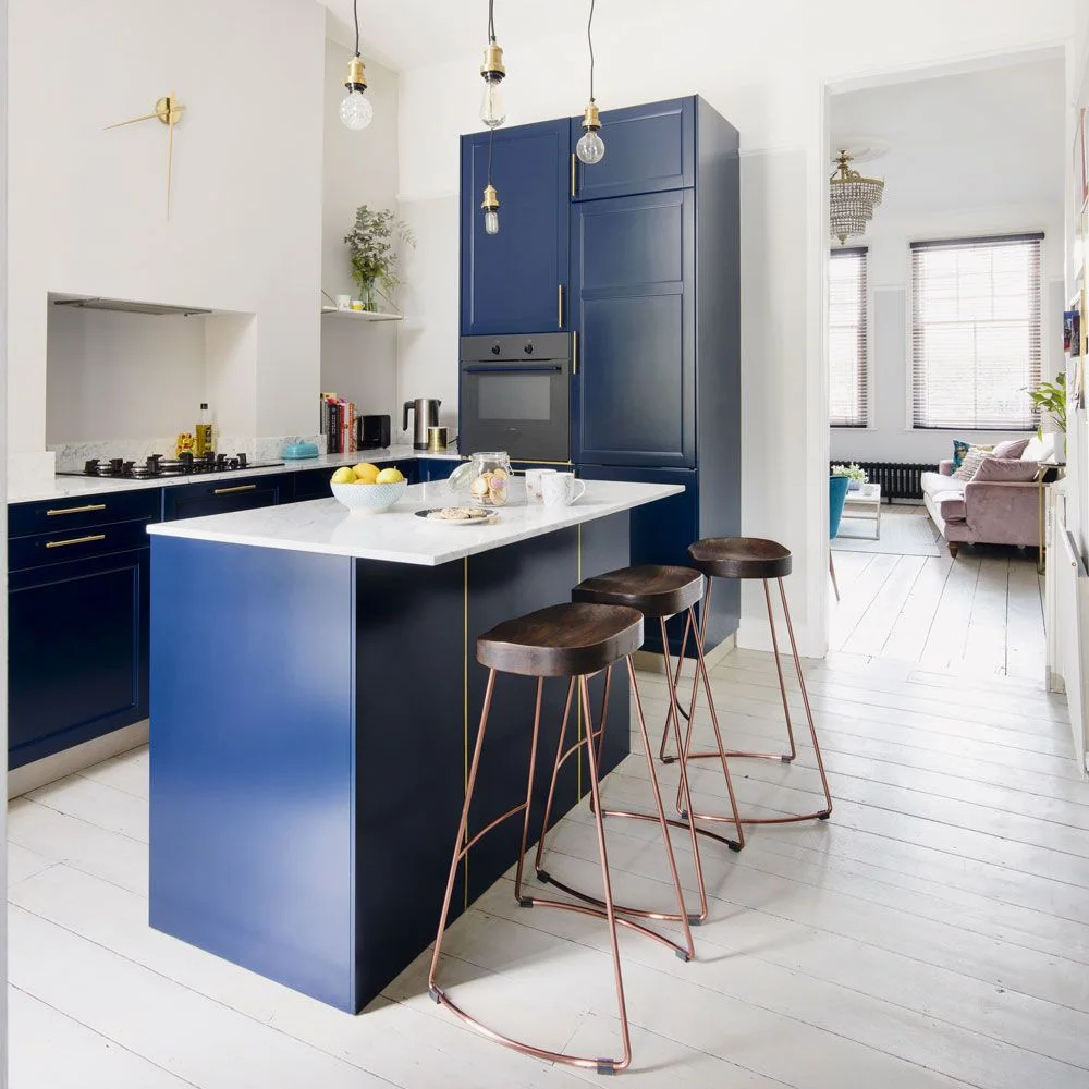 Navy kitchen cabinets and island with a white counter, wooden stool seats, warm-metal handles and white floorboards.