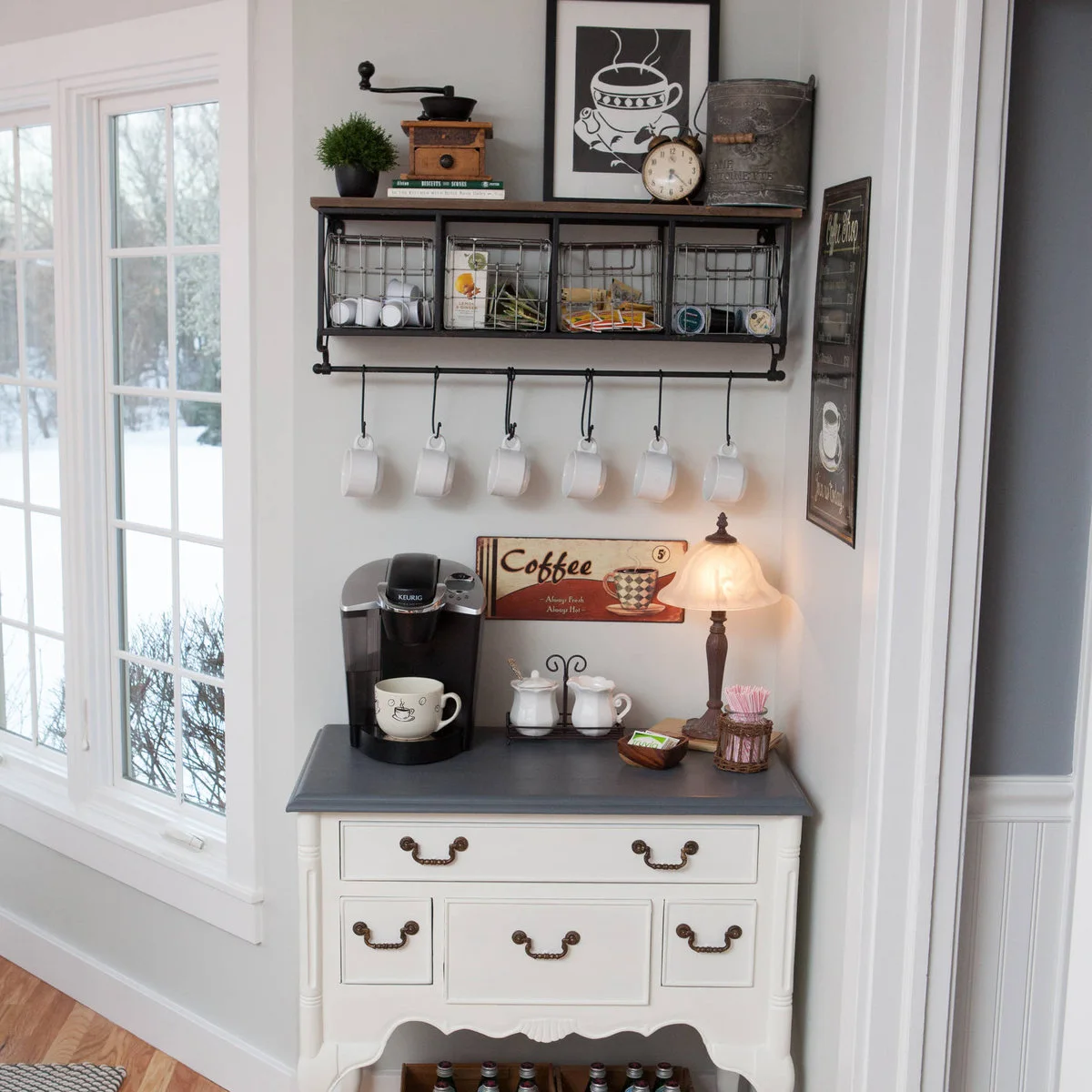 Small white chest with coffee maker beneath a wall rack of wire baskets and hanging mugs