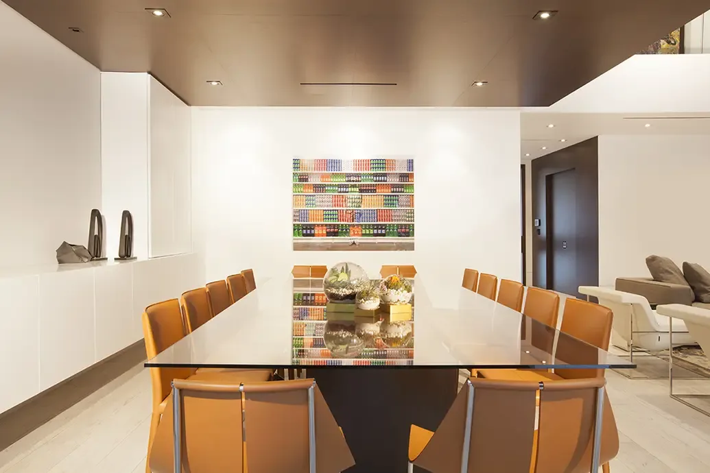 Dark glass dining table with orange chairs and a dark central base beneath a brown ceiling beside colorful framed art
