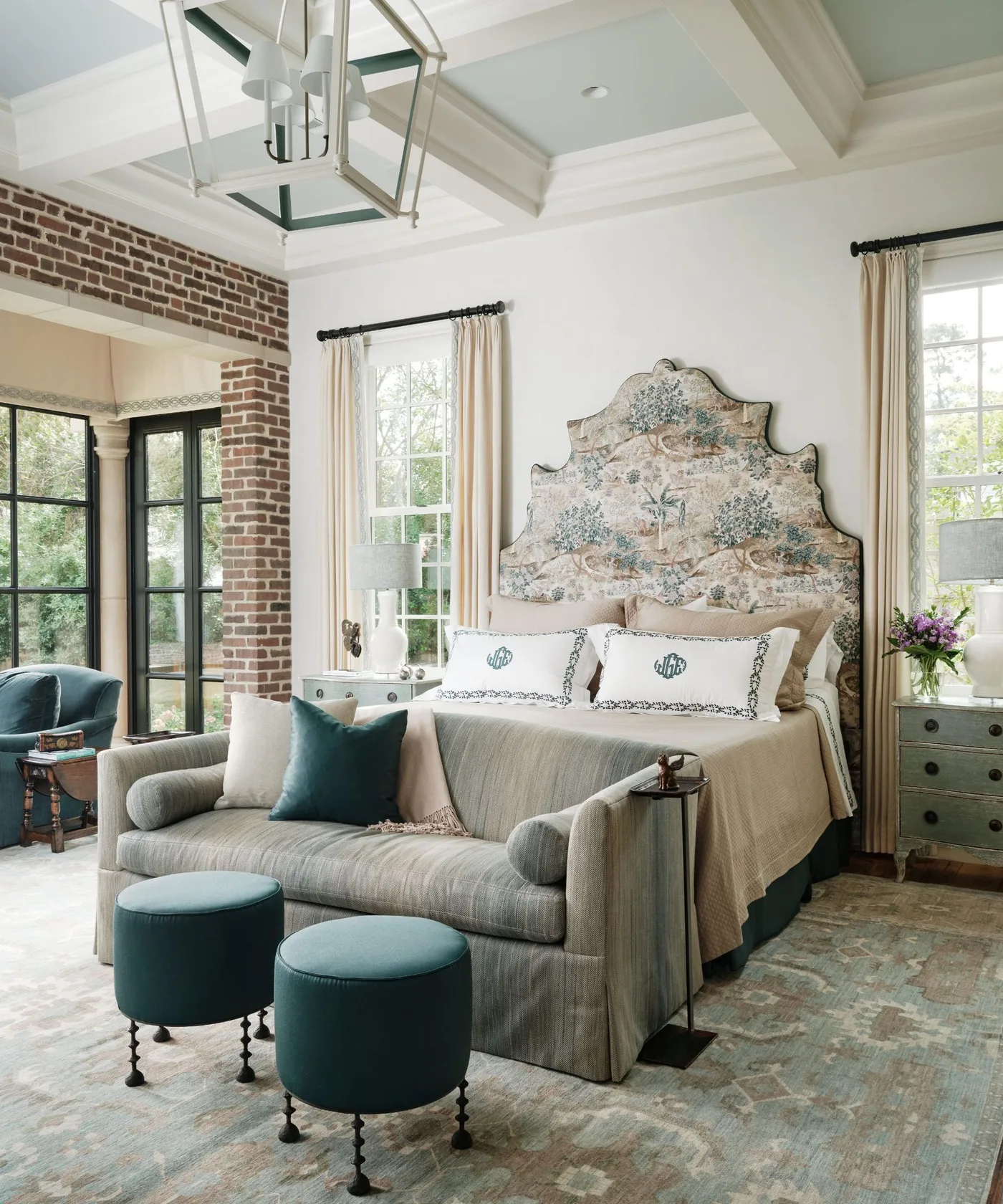 Country bedroom with a tall shaped patterned headboard, pale bedding, striped settee at the bed foot, dark green stools and long cream curtains.