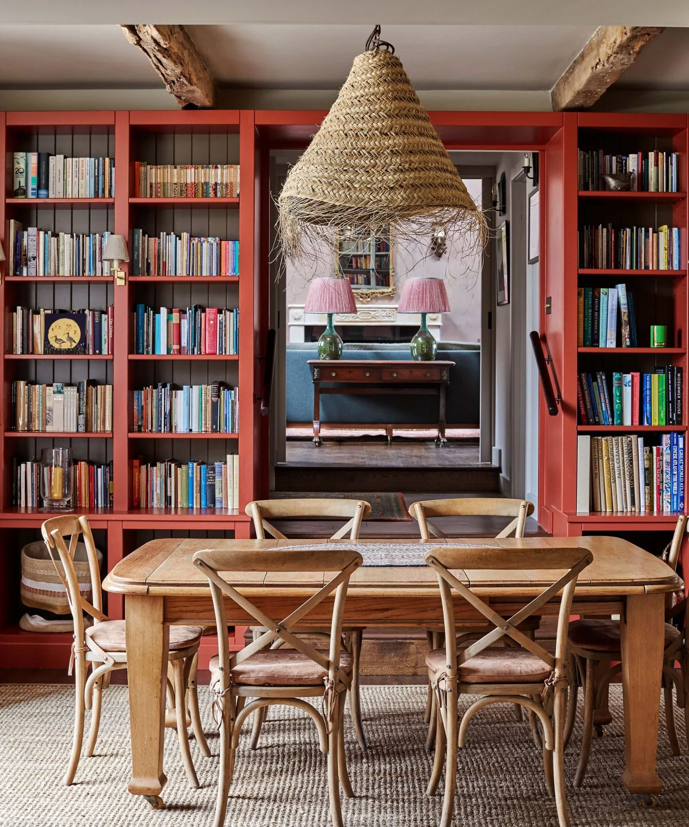 Red bookshelves around a dining doorway behind a wood table, cross-back chairs and a woven pendant
