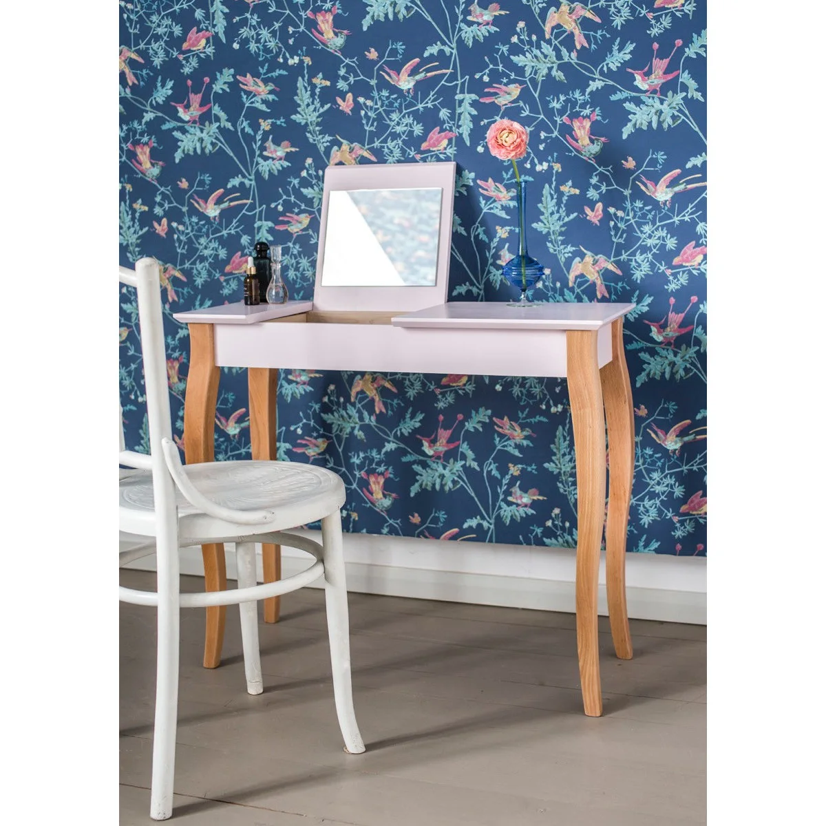A pale pink dressing table with timber legs and a raised mirror section stands against blue botanical wallpaper