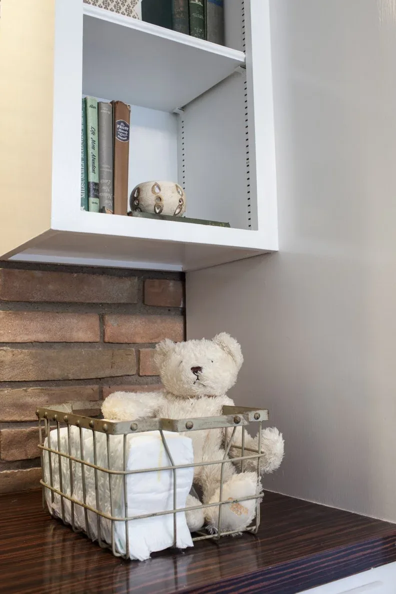 Wire basket holding folded white cloths and teddy bear on dark wood surface beneath white shelves and brick wall