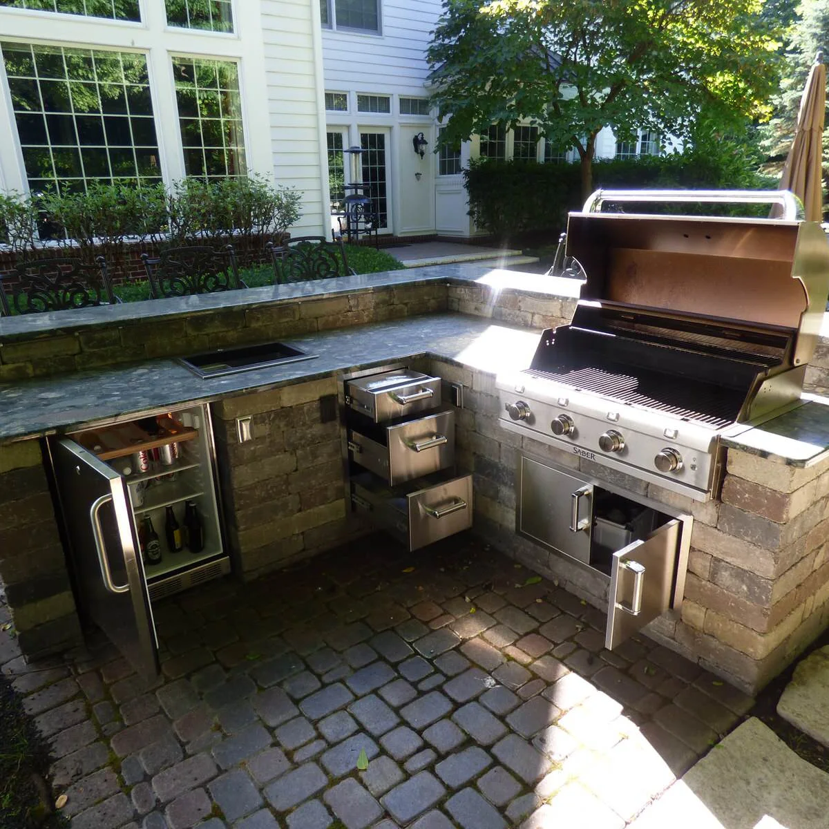L-shaped outdoor grill kitchen showing open drawers, cabinet door and beverage refrigerator door over a paved floor