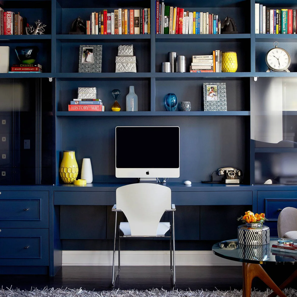 A blue built-in desk with a computer beneath bookshelves, beside an armchair and glass coffee table.